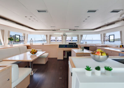 A spacious and modern interior of the Lagoon 560 catamaran. The salon features comfortable seating, a dining table, and large windows offering stunning sea views. The well-equipped kitchen is perfect for preparing delicious meals.