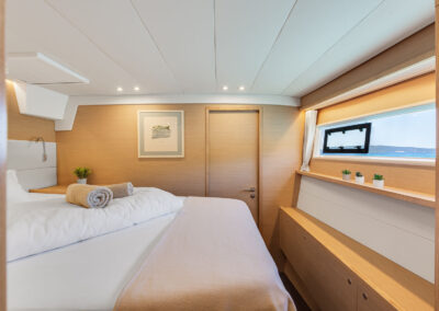 A luxurious bedroom on the Lagoon 560 s2 catamaran, with a king-size bed, wooden furnishings, and a large window with a blue sea and islands view.