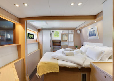 A luxurious bedroom on the Lagoon 560 s2 catamaran, with a king-size bed, wooden furnishings, and a large windows letting in the natural light. Island's greenery is seen through the windows.