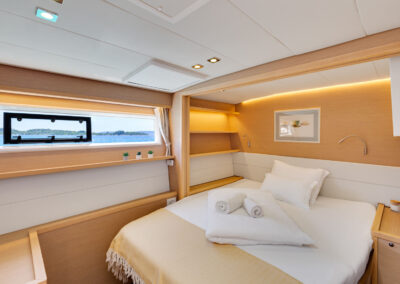 A luxurious bedroom on the Lagoon 560 s2 catamaran, with a king-size bed, wooden furnishings, and a large window with a blue sea and islands view.