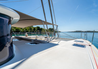 A stunning view of the Lagoon 560 catamaran's spacious aft deck. The deck features comfortable shaded seating area, a small table, and 2 sunbathing nets. In the background, you can see a breathtaking view of the Croatian coastline and crystal-clear sea.