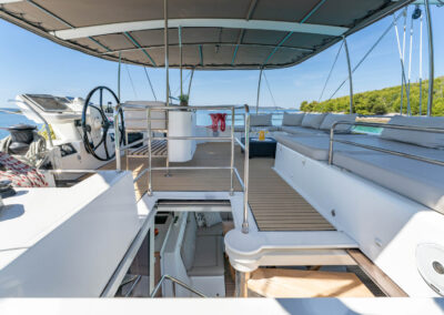 A stunning view of the Lagoon 560 catamaran's spacious flybridge. The deck features comfortable seating, a dining table, and a helm station. In the background, you can see a breathtaking view of the Croatian coastline and crystal-clear sea.