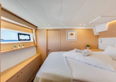 A luxurious bedroom on the Lagoon 560 s2 catamaran, with a king-size bed, wooden furnishings, and a large window with a blue sea and islands view.