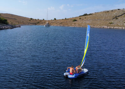 A couple sailing a small inflatable sailboat in a calm bay with a catamaran in the background.