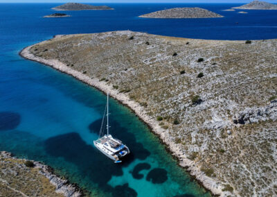 A stunning aerial view of a Lagoon 560 catamaran anchored in a secluded bay on the Croatian coast. The crystal-clear waters, rocky islands, and picturesque landscape create a breathtaking scene.