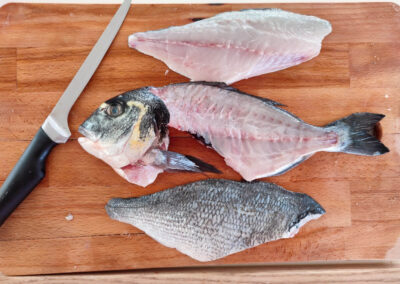 A fresh fish, possibly a sea bream, is being filleted on a wooden cutting board. The fish is surrounded by its fillets, showcasing the freshness and quality of the ingredients used in our onboard cuisine.