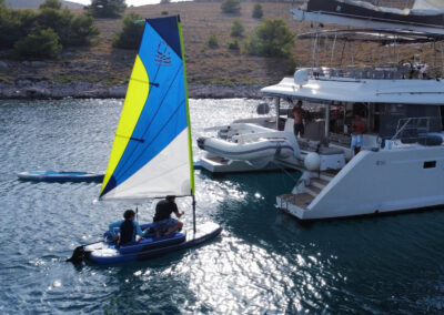 A family enjoying water sports, with a small sailboat and paddleboard in front of a luxury catamaran.